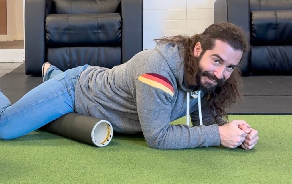 Dr. John is lying on his stomach with the foam roller under his right leg. Using his arms to hold himself up, he is rolling the inside of his groin and adductor muscle